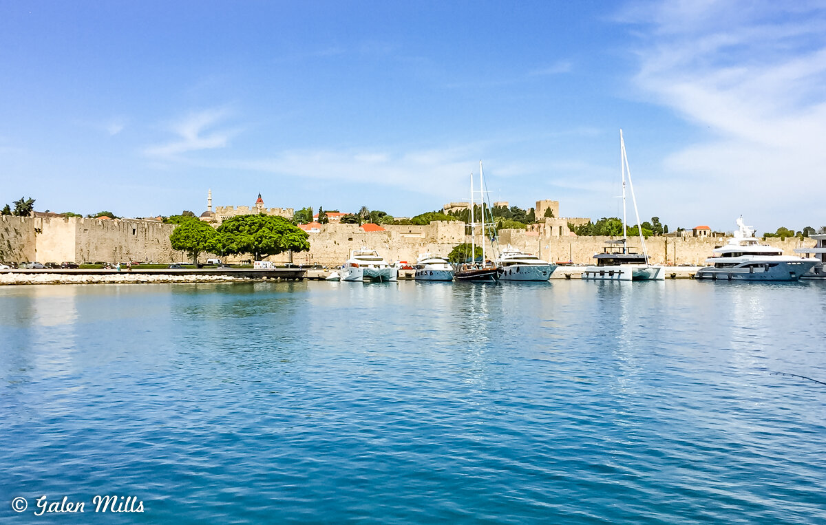 View of Rhodes Marina with boats and medieval city walls, clear blue sky, and calm water, creating a picturesque scene.