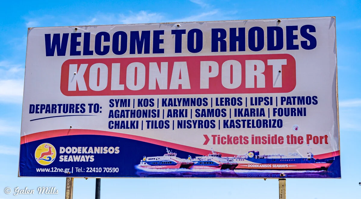 Sign reading 'Welcome to Rhodes, Kolona Port,' listing ferry departures to various Greek islands.