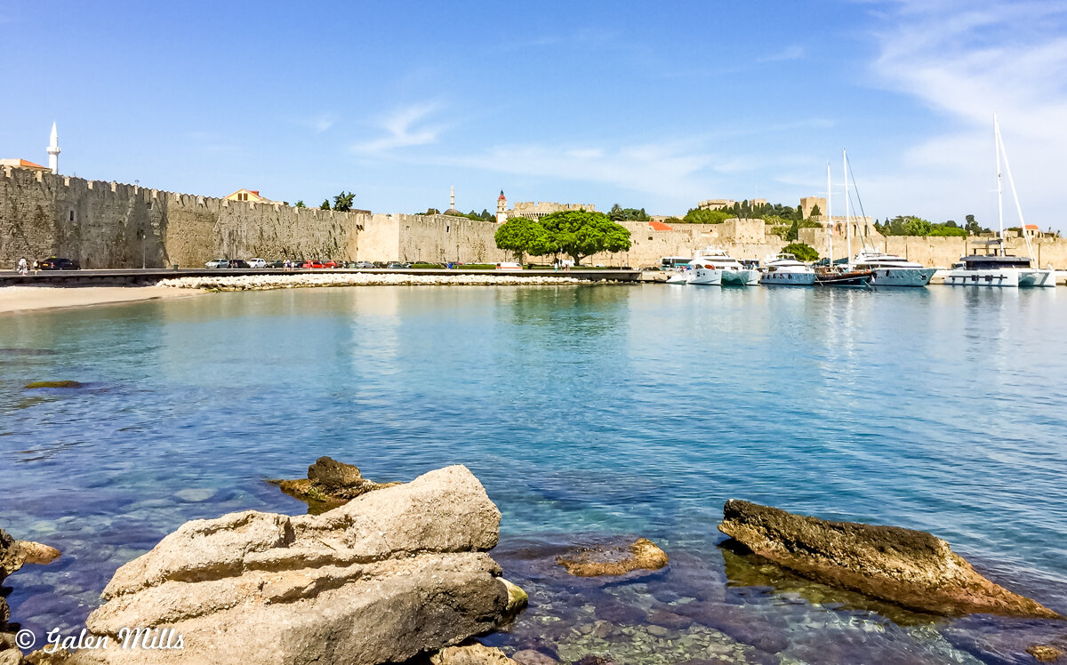 Coastal view with fortress walls, small beach, docked boats, and clear blue water