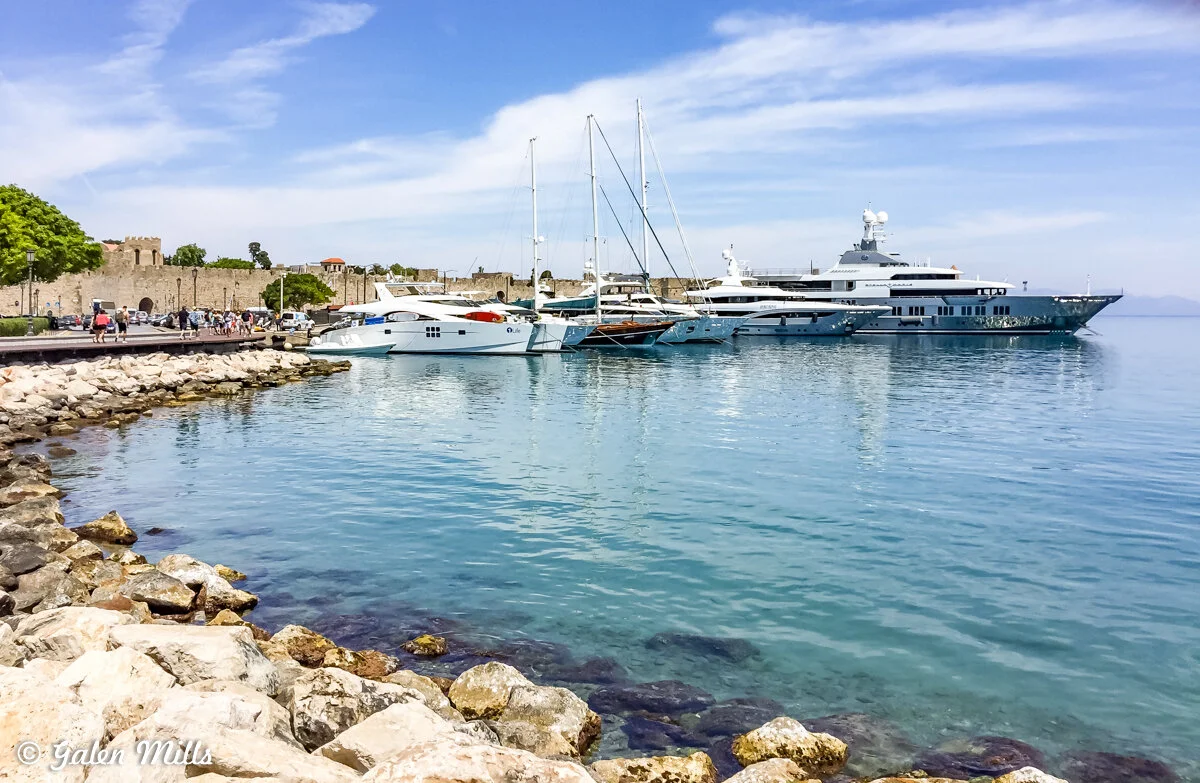 Marina with yachts and boats docked along the waterfront on a sunny day, with rocky shoreline and historic buildings in the background.