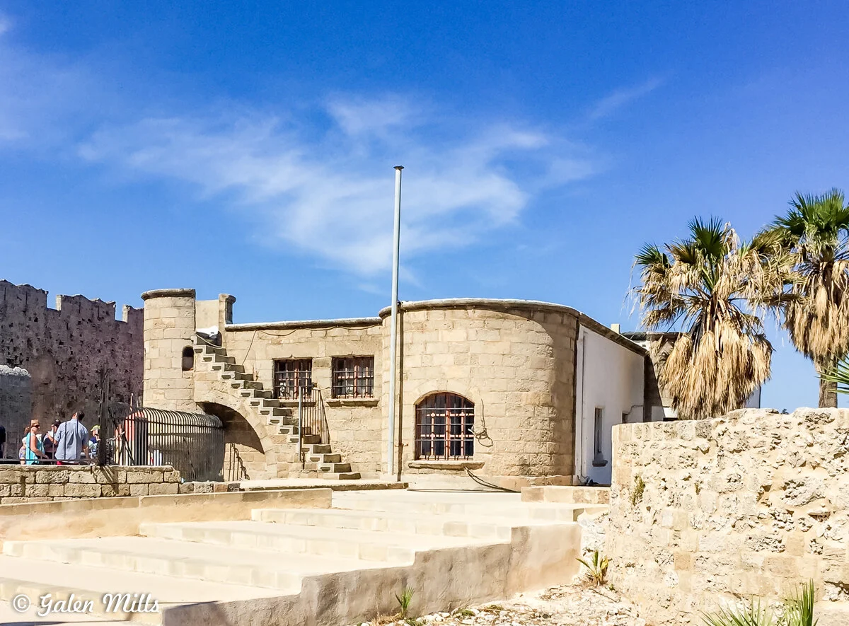 Historic stone building with arched windows and external staircase, surrounded by palm trees and blue sky.