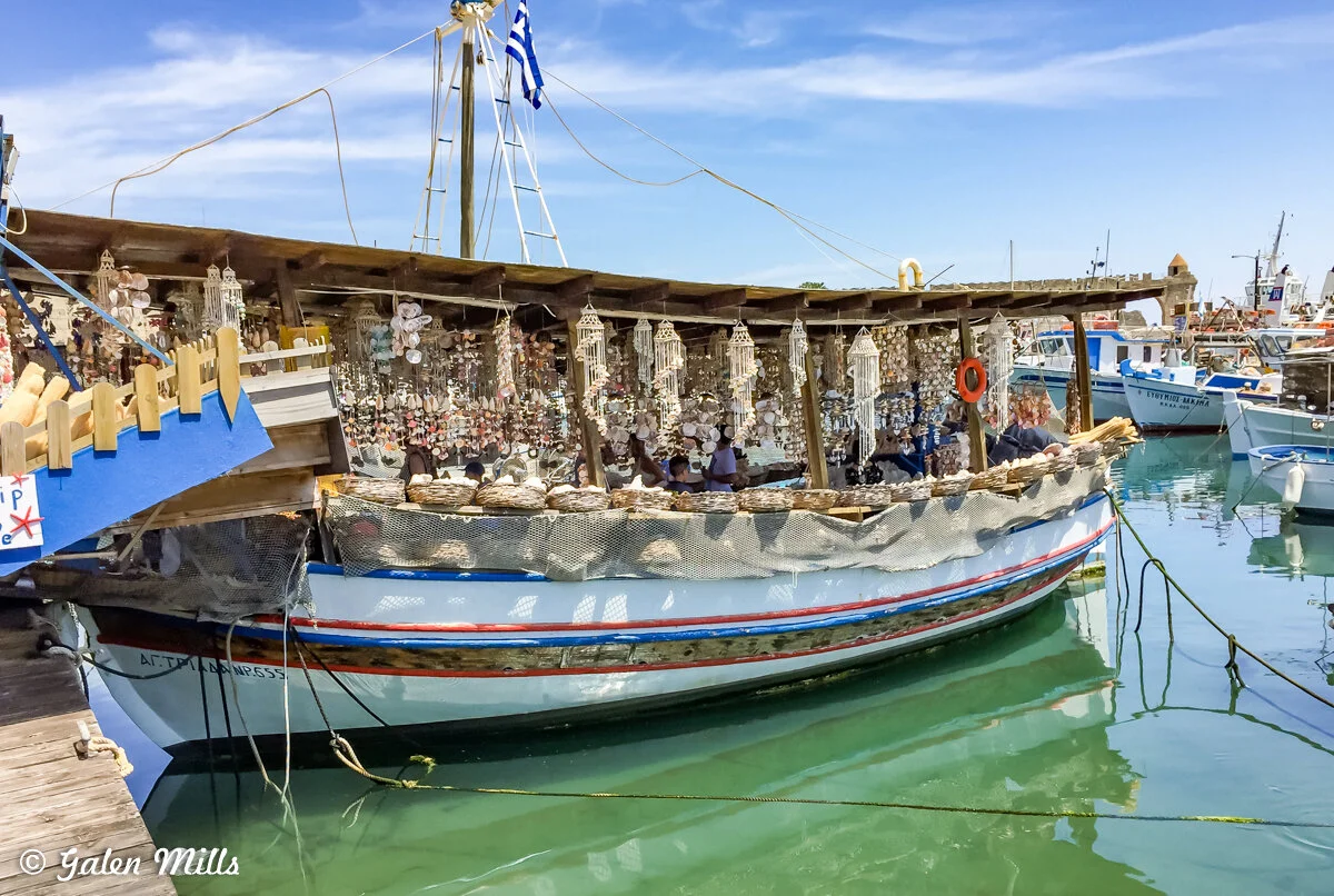 Traditional Greek boat in a harbor decorated with hanging shells and crafts, with a Greek flag.