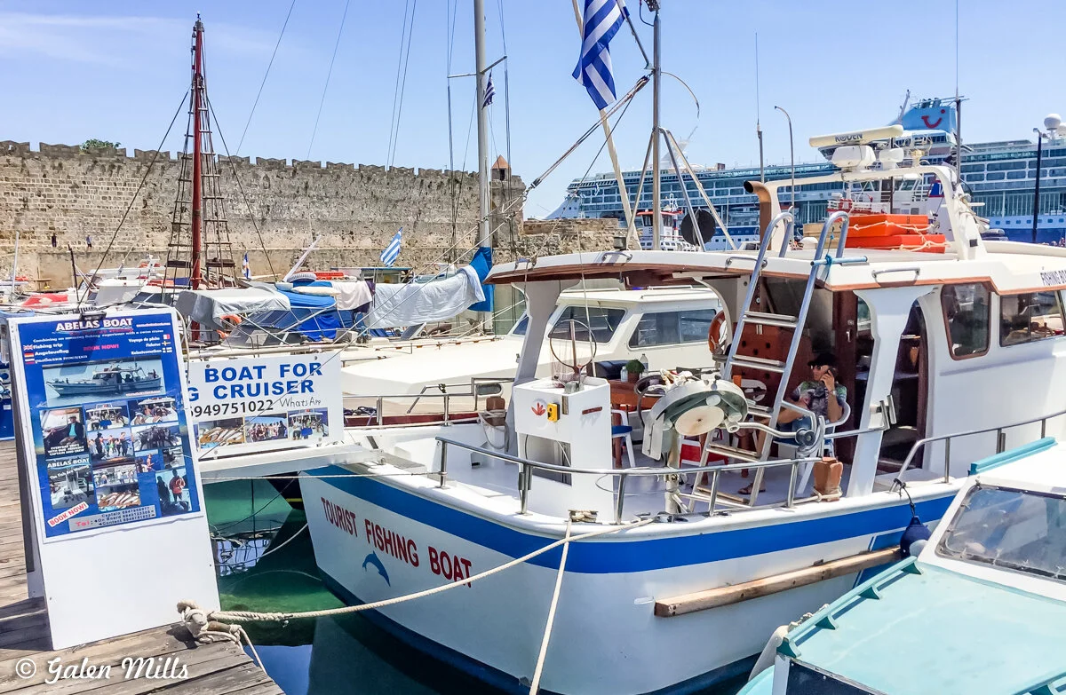 Boats docked in a harbor with a tourist fishing boat prominently labeled. A sign promotes boat tours with images and contact information. A stone wall and a large cruise ship are visible in the background.