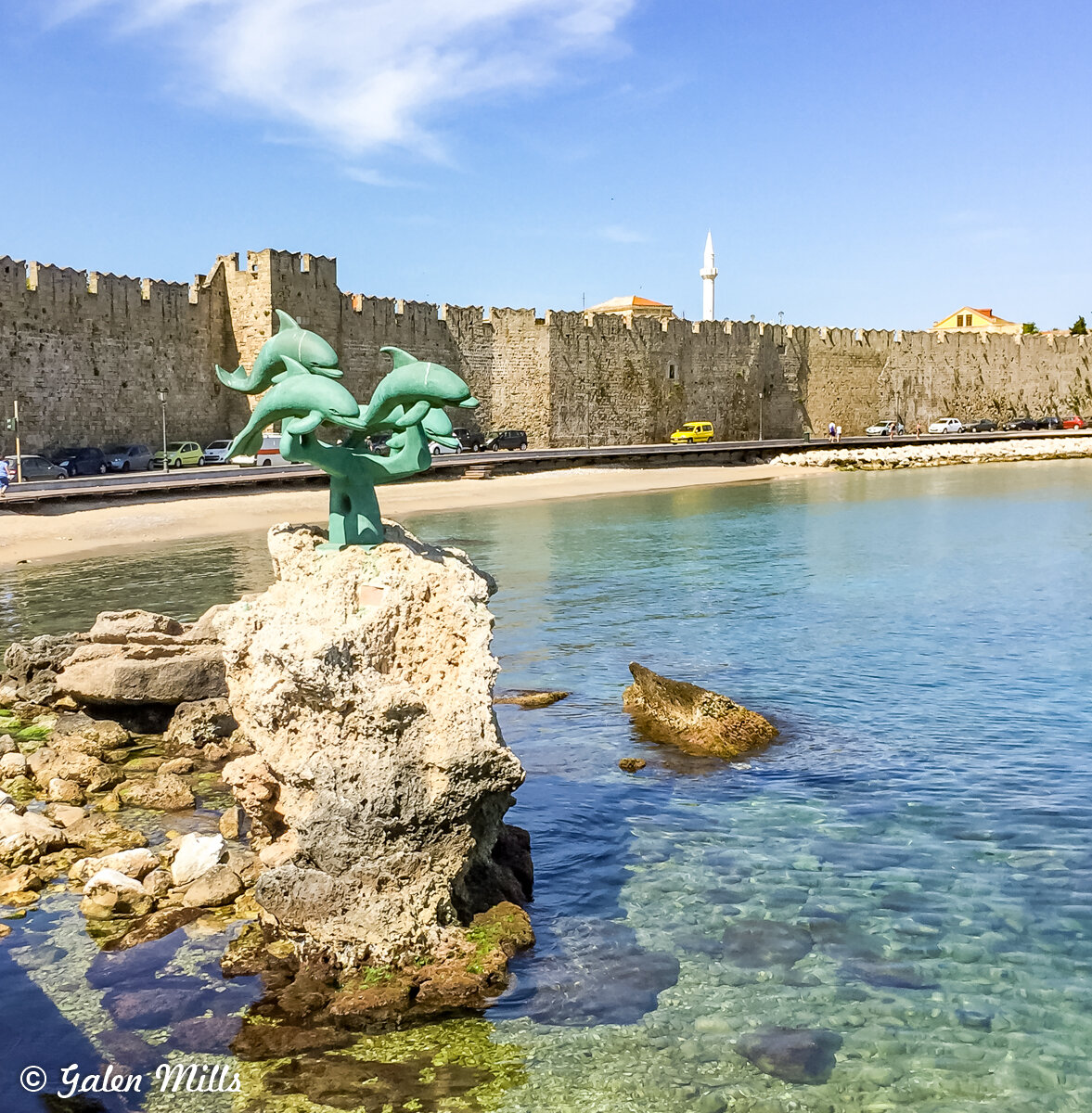 A sculpture of dolphins on a rock in a harbor with a medieval stone wall and buildings in the background.
