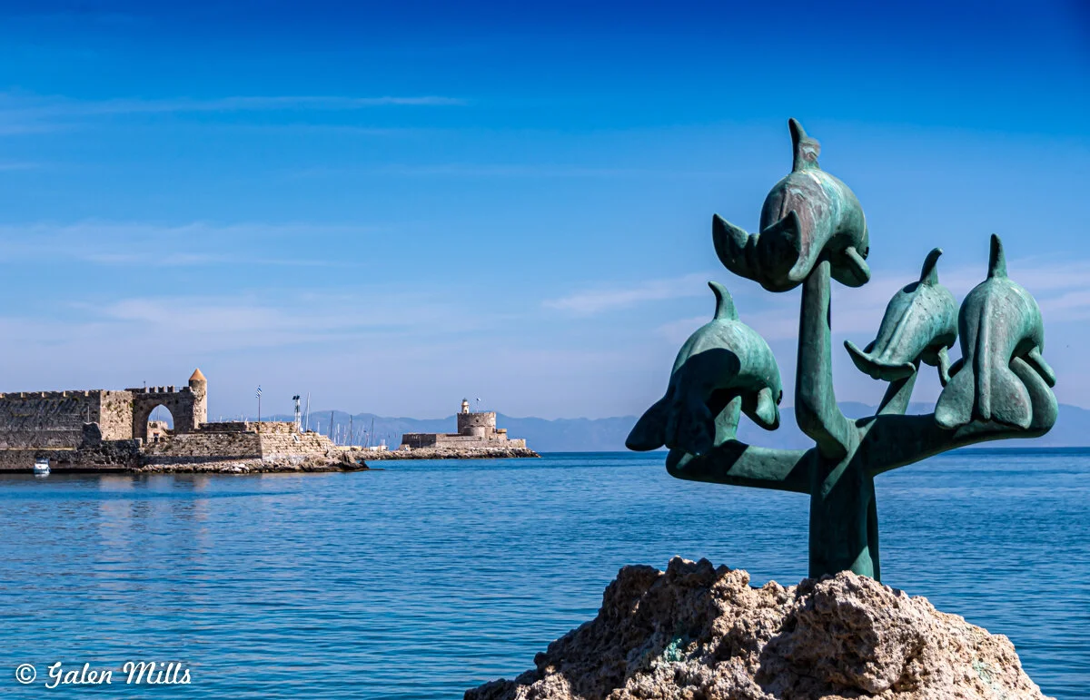 Bronze dolphin sculpture near a harbor in Rhodes, Greece with medieval fortifications and a lighthouse in the background under a clear blue sky.