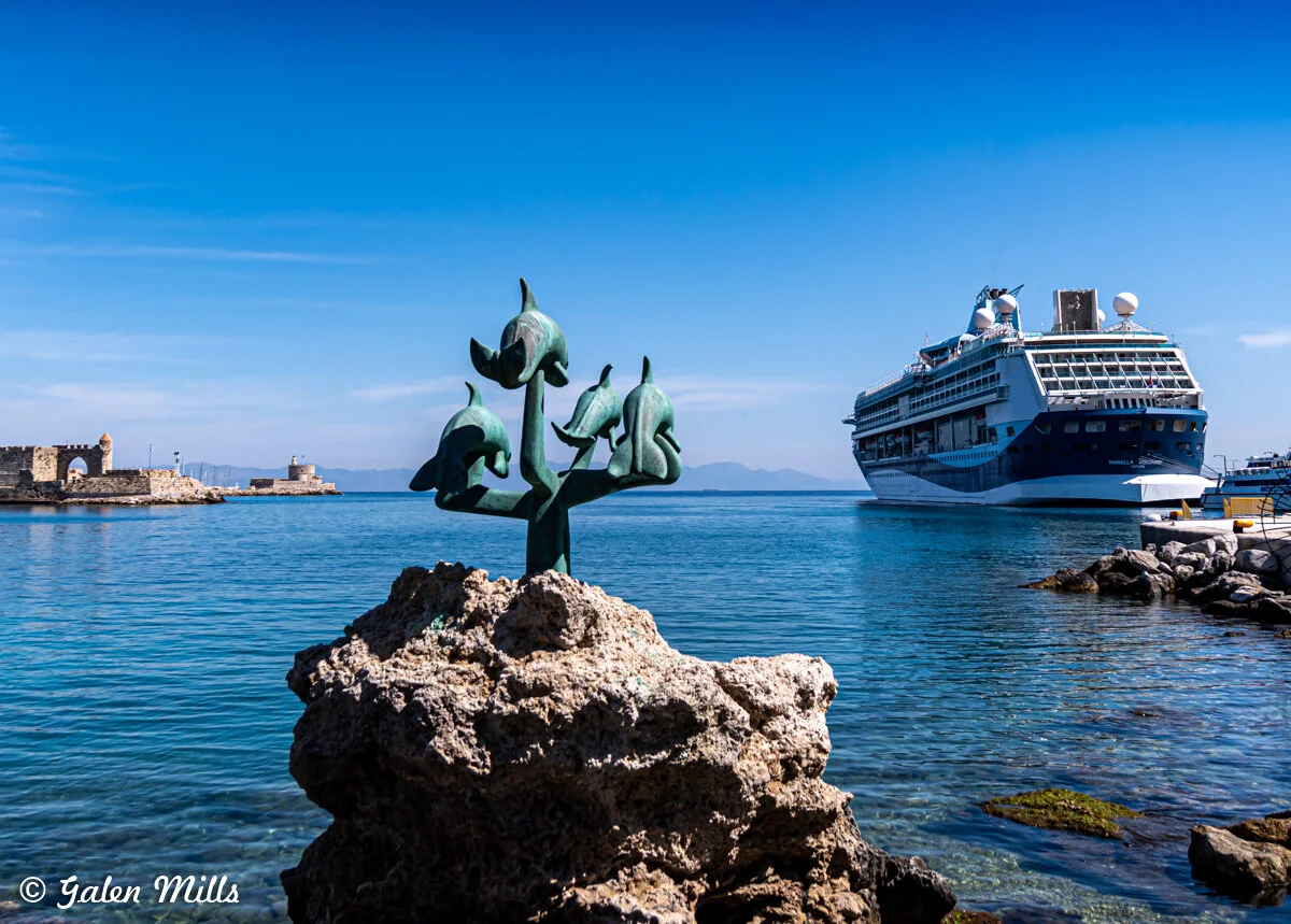 Sculpture of dolphins on a rock near the sea with a cruise ship and fortress in the background.