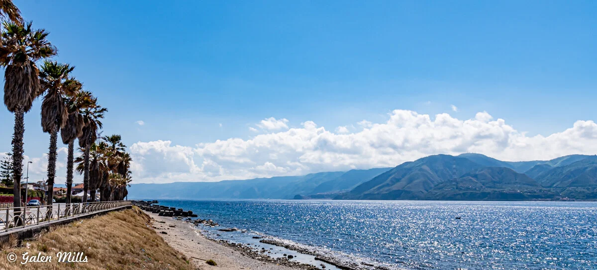Coastal landscape with palm trees, ocean, and mountains under a clear blue sky.