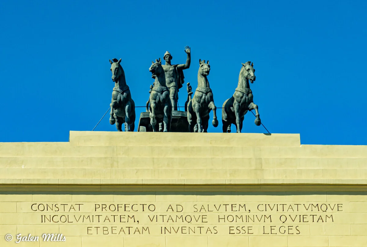Stone sculpture of a chariot with four horses and a driver on top of a building, featuring Latin inscription below, against a clear blue sky.