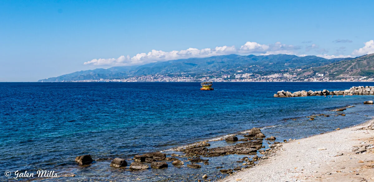Coastal scene with blue ocean, rocky shoreline, distant city, green hills, and cloudy sky.