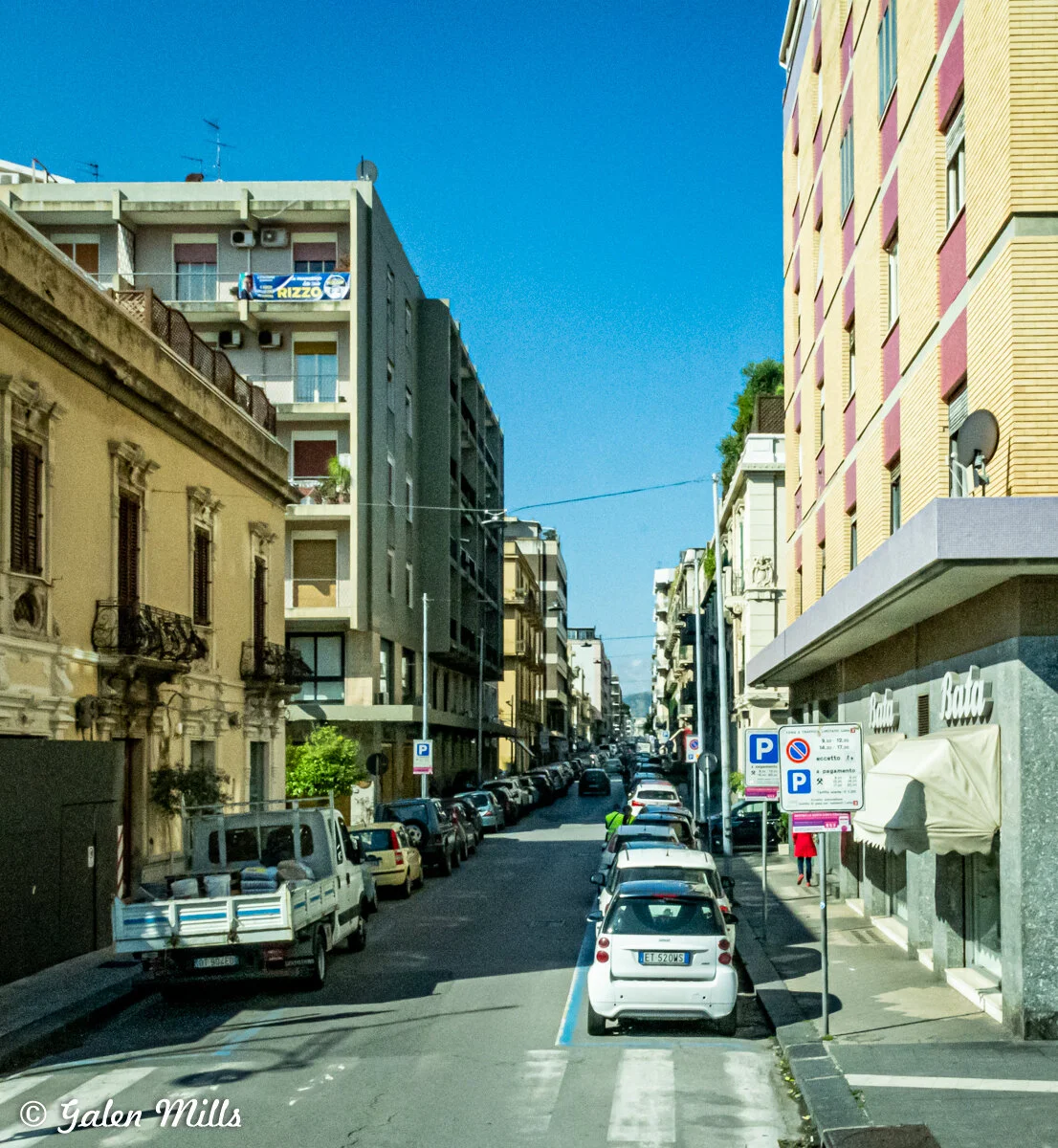 City street with parked cars and apartment buildings in an urban area under a clear blue sky.