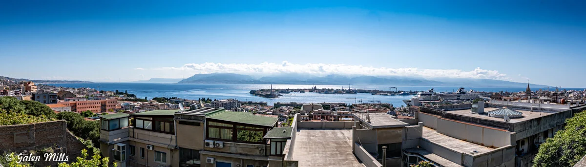 Panoramic view of a coastal city with buildings, rooftops, a harbor, and distant mountains under a clear blue sky.