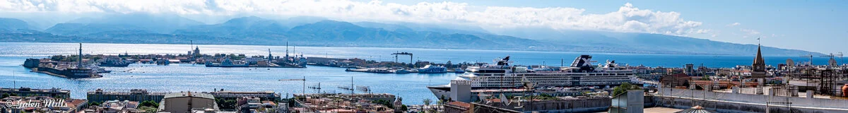 Panoramic view of a coastal city with a harbor, featuring buildings, docks, ships, and distant mountains under a cloudy sky.