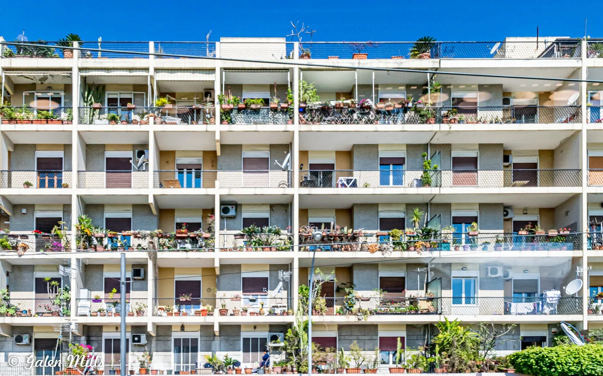 Exterior of an apartment building with multiple balconies decorated with plants and furniture under a blue sky.