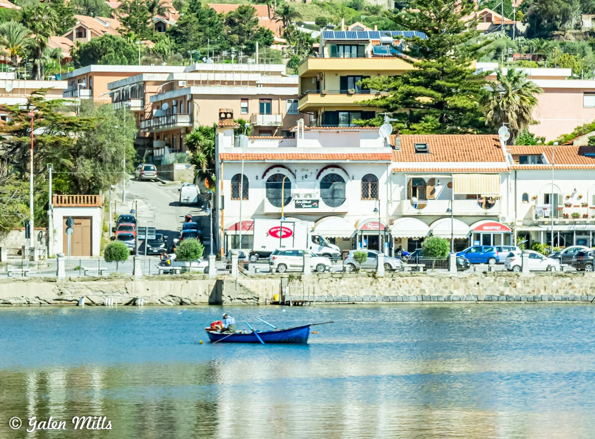 Scenic view of a small boat on a calm river with an urban backdrop, featuring buildings with red-tiled roofs and parked vehicles along the waterfront.
