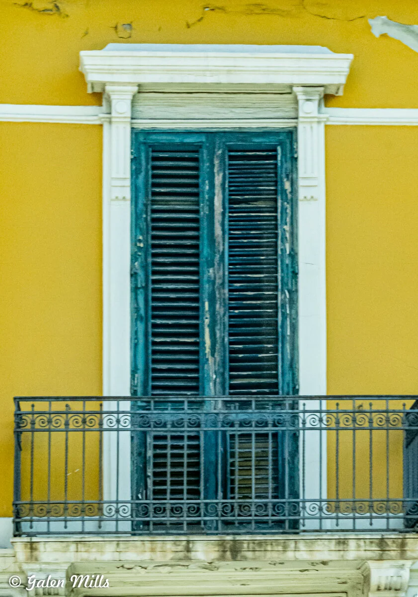 A weathered blue wooden shuttered window with ornate black iron balcony railing against a yellow wall.