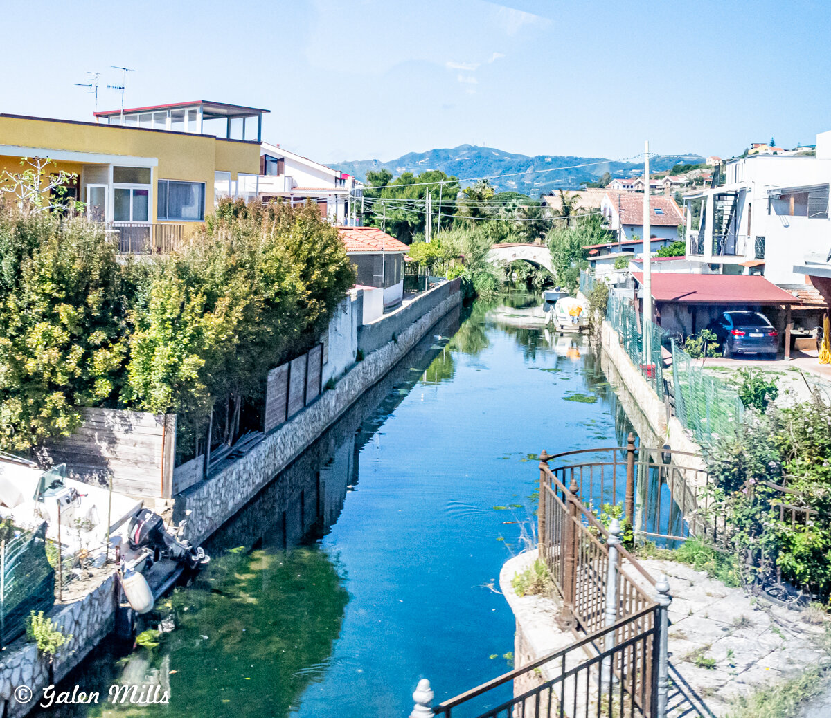 Narrow canal with clear blue water flanked by residential houses and greenery, featuring a boat docked on one side, with distant mountains and a clear sky in the background.
