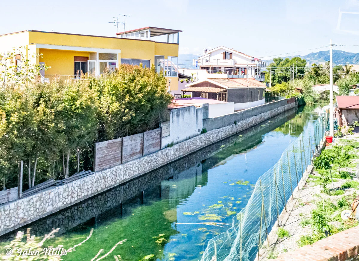 Canal bordered by residential homes with yellow buildings and greenery in a bright neighborhood setting.