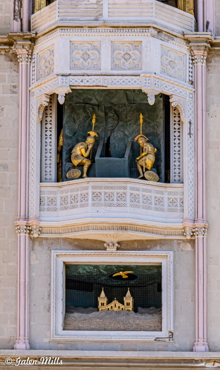 Decorative building facade with statues of Roman soldiers and a scene depicting a bird above a church.