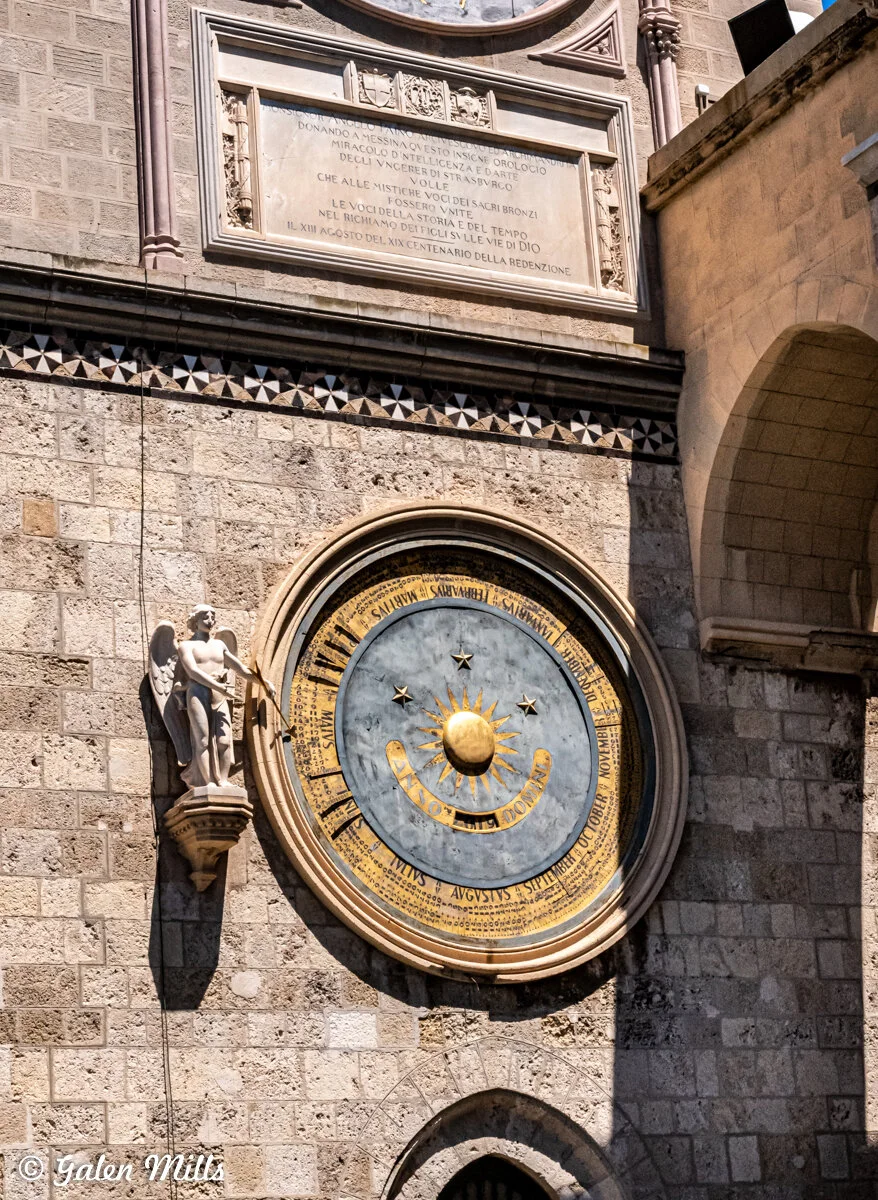 Ancient astronomical clock on a stone wall with a statue of an angel and sunlight casting shadows.