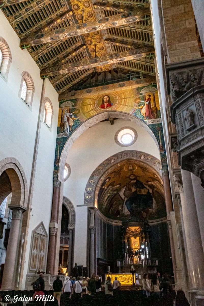 Interior of a church with intricate ceiling designs, arches, religious mosaics, and a group of people walking. The artwork features religious figures and gold accents.
