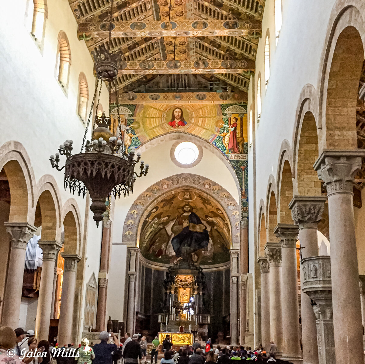 Interior of a historic church with ornate ceiling, chandeliers, arched columns, religious paintings, and an altar, populated by visitors and tourists.