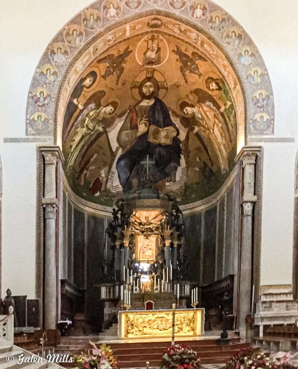 Interior of an ornate church altar with religious mural and statue, featuring candles and floral arrangements.