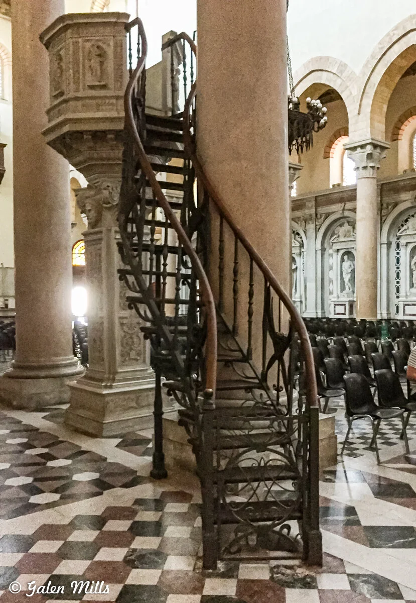 Ornate spiral staircase inside a cathedral with stone columns and arched windows