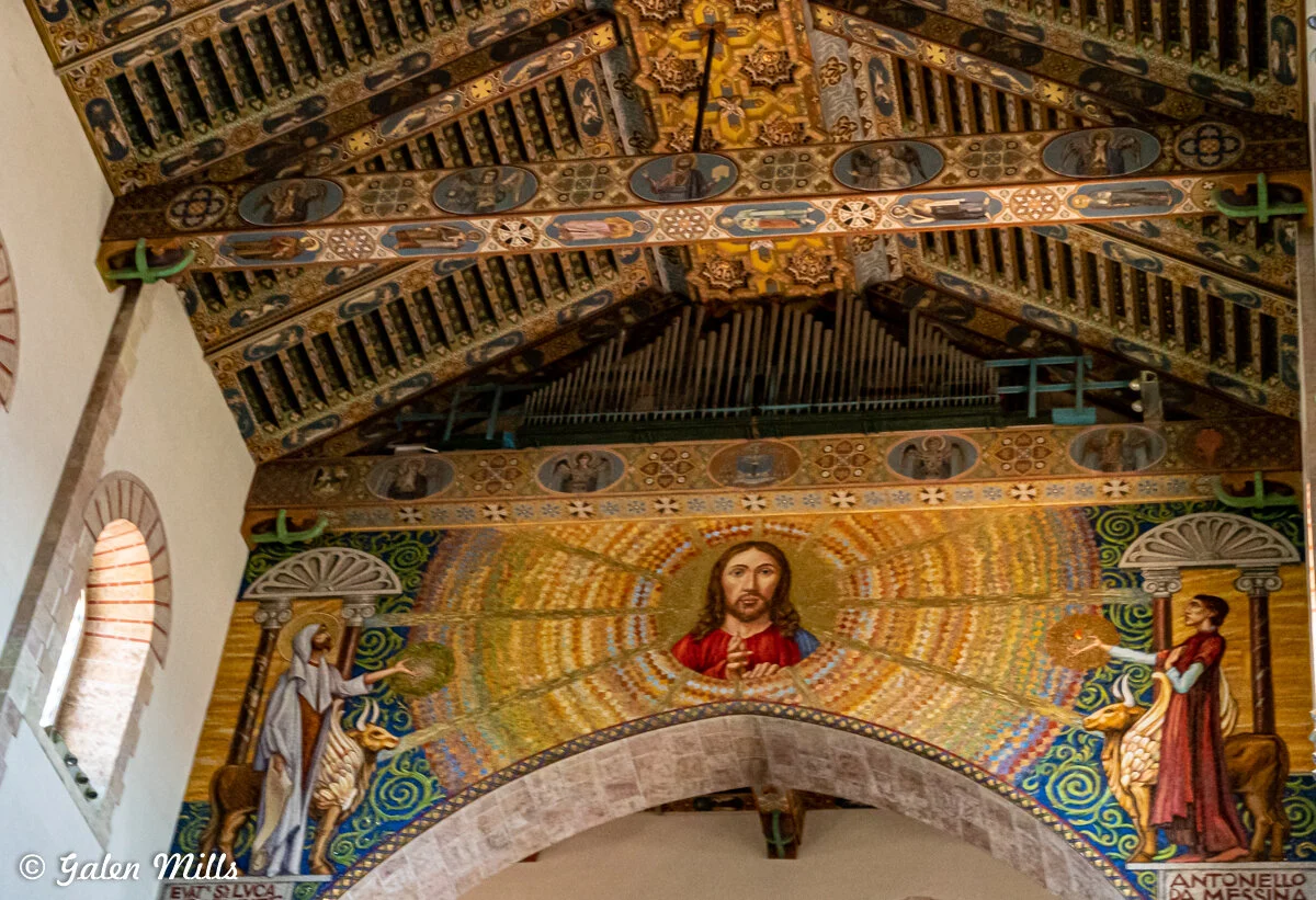 Decorative church ceiling featuring a central religious figure in a radiant setting, flanked by two other figures, with ornate patterns and an organ above.
