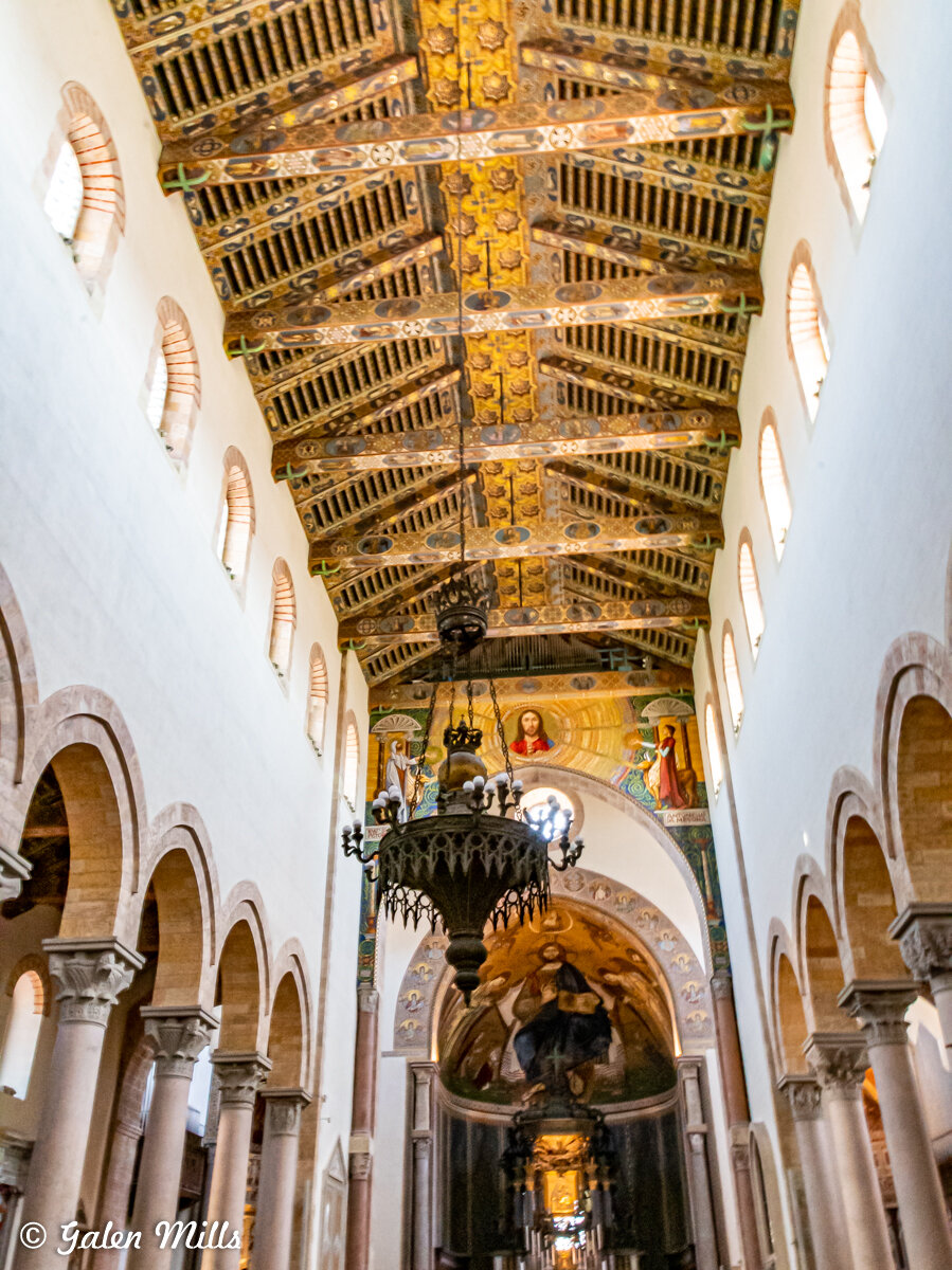 Interior of Monreale Cathedral in Sicily, featuring ornate wooden ceiling, large chandelier, and arches.