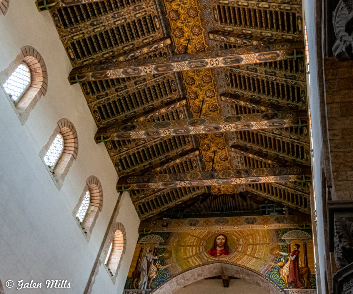 Interior of a church featuring a detailed wooden ceiling with ornate patterns, decorative beams, and religious artwork depicting figures and symbols on the arch. Arched windows with stone accents line the wall.