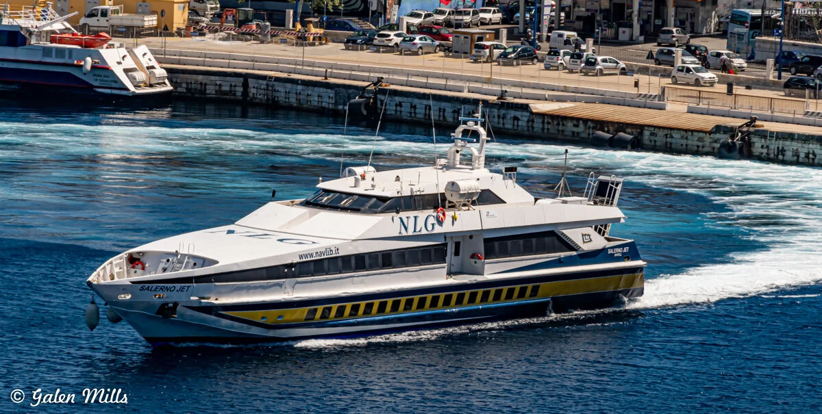 A white high-speed ferry named Salerno Jet sailing near a harbor with docking facilities and parked cars in the background.