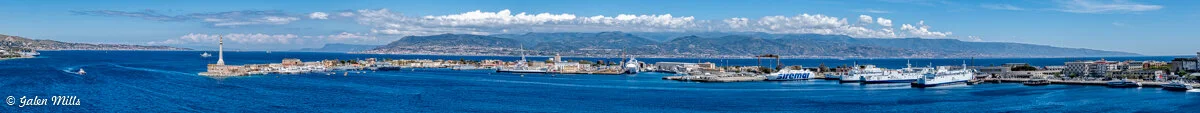 Panoramic view of a coastal cityscape with a port, lighthouse, and ferries, surrounded by mountains and clear blue sea.