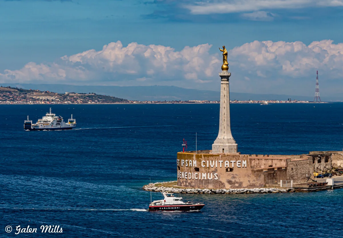 A coastal scene with a tall lighthouse-like structure featuring a golden statue on top. It is situated on a stone fortification in the sea. The phrase "ET IPSAM CIVITATEM BENEDICIMUS" is inscribed on the structure. Nearby, a carabinieri boat and a fe