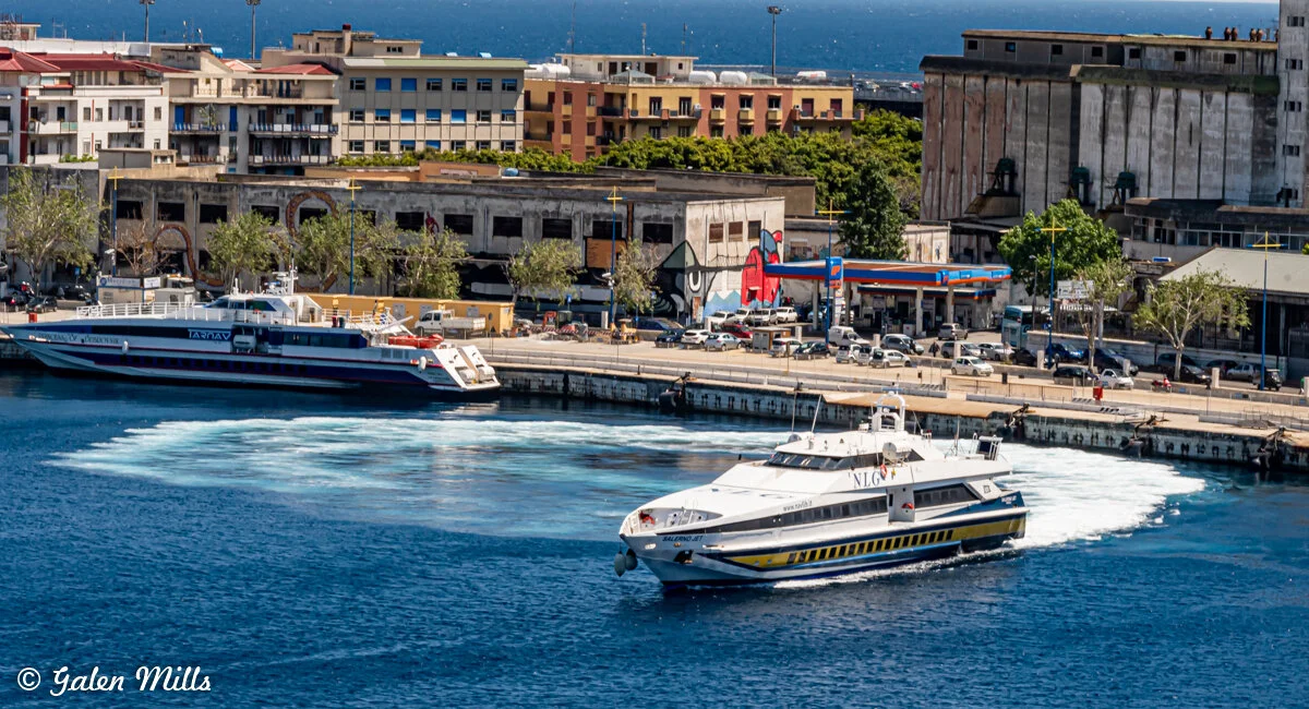 Two ferries docked at a harbor with a cityscape background, featuring buildings and a mural on the wall, under a clear blue sky.