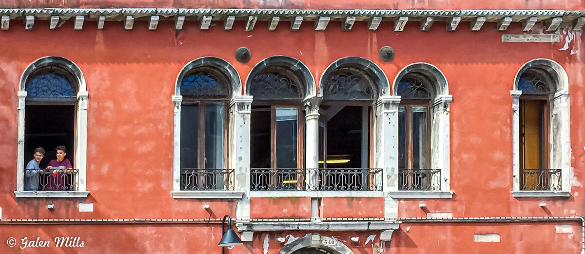 Facade of a historic building in Venice with arched windows and balcony railings. Two people stand looking out from a window on the left. The wall is painted orange-red with weathered patches.