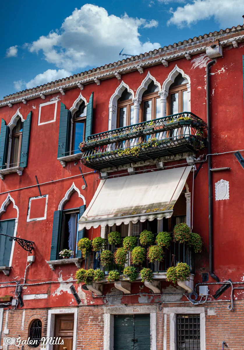 Old red building with ornate white-framed windows, green shutters, and balconies with plants against a blue sky.