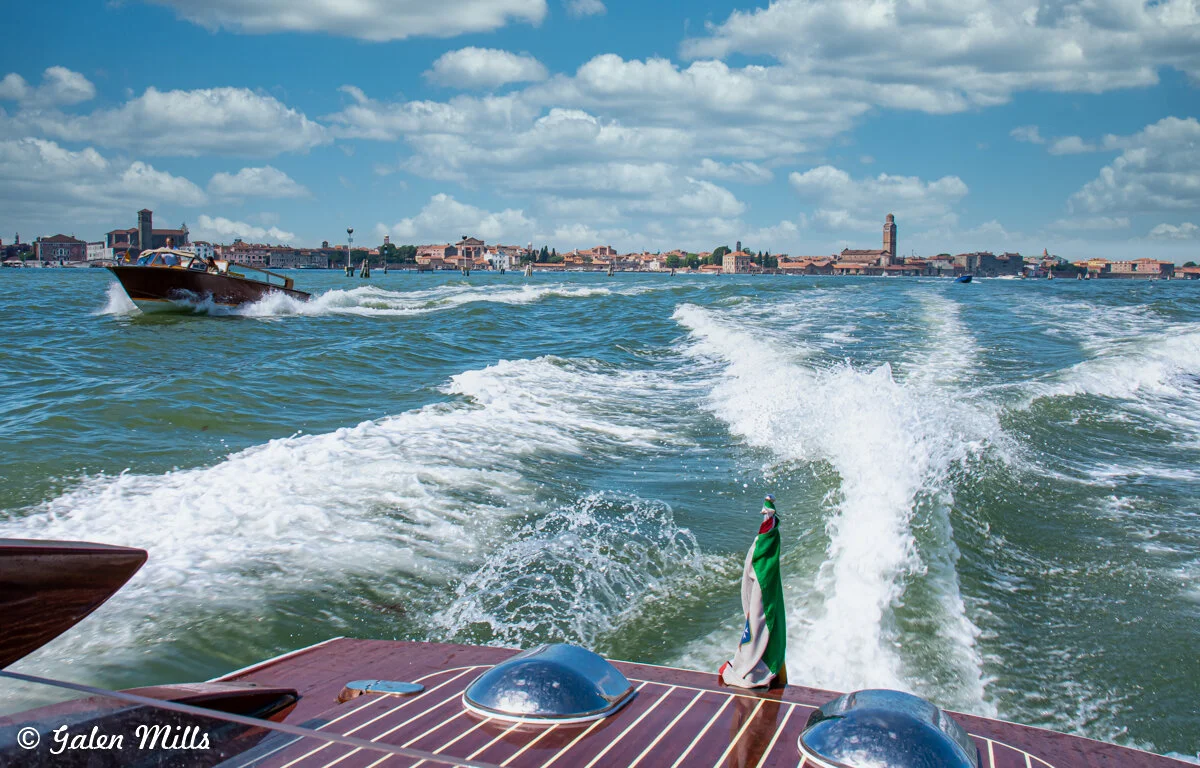 Wooden boat moving on a waterway near a coastal city with another boat in view, clear blue sky with scattered clouds.