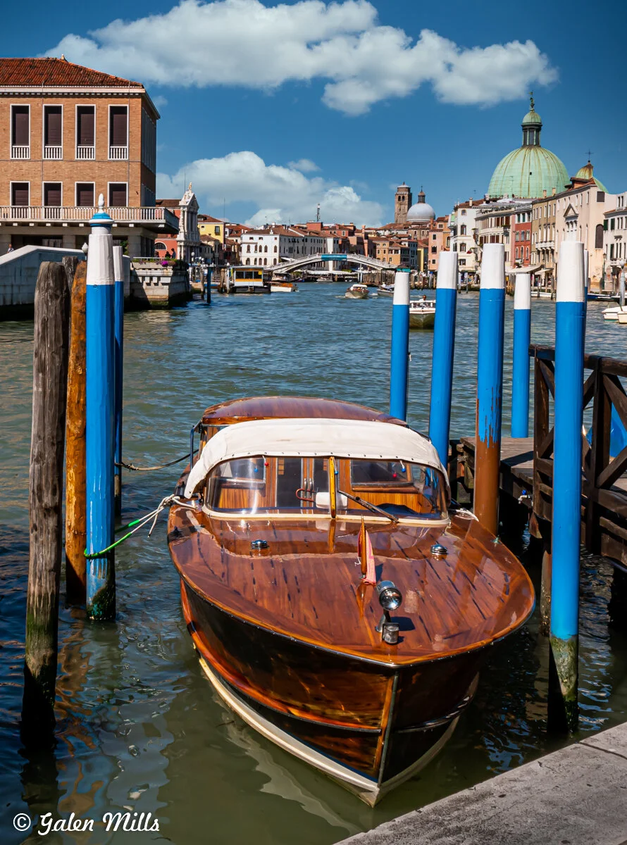 Wooden boat docked on a canal in Venice, Italy, with colorful buildings and San Simeone Piccolo in the background.
