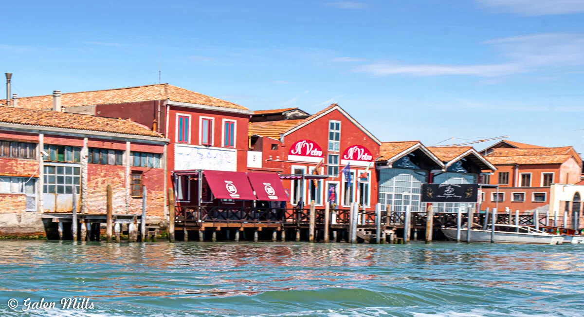 Colorful buildings along a canal in Murano, Italy, with brick facades and red awnings reflecting on the water.