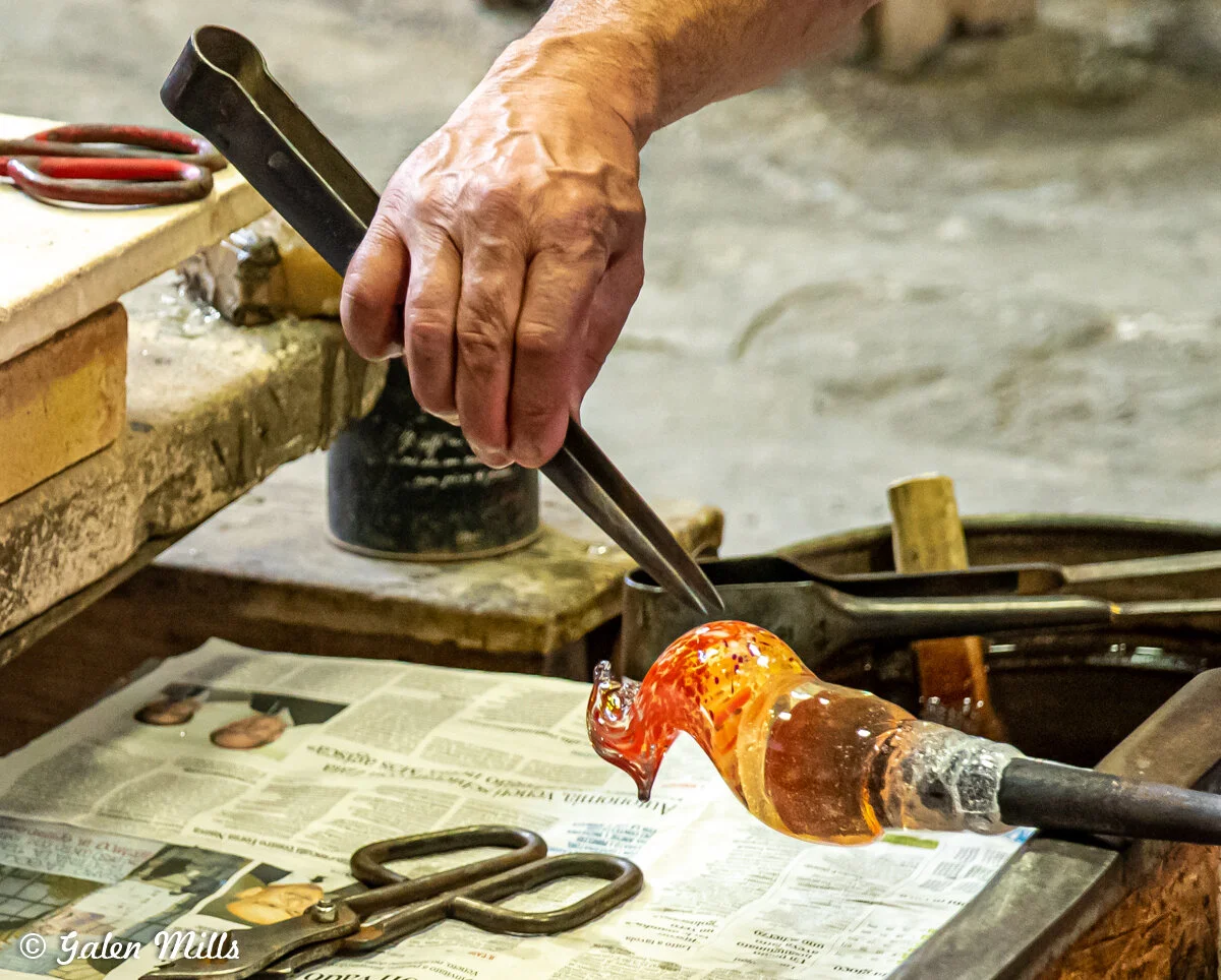 Glassblower shaping hot glass with tongs on workbench covered with newspaper.