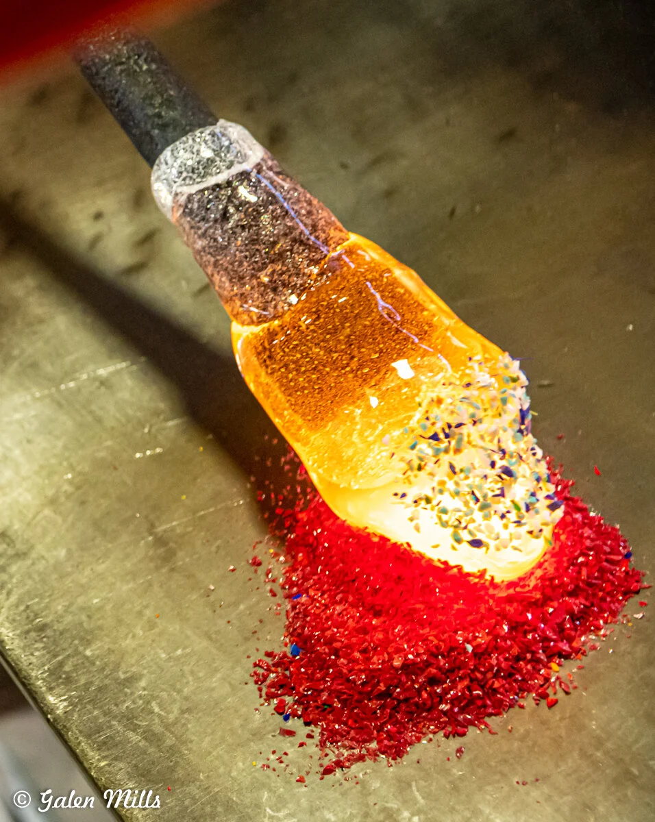 Glassblowing process with a hot glass piece on a rod being rolled on colored glass frit, creating a vibrant design.
