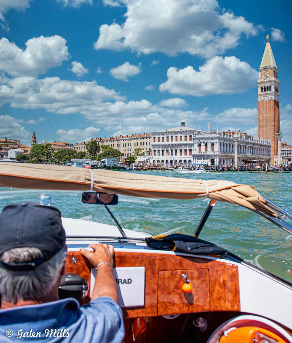 Person steering a boat on a canal in Venice, Italy with historic buildings and St. Mark's Campanile visible in the background.