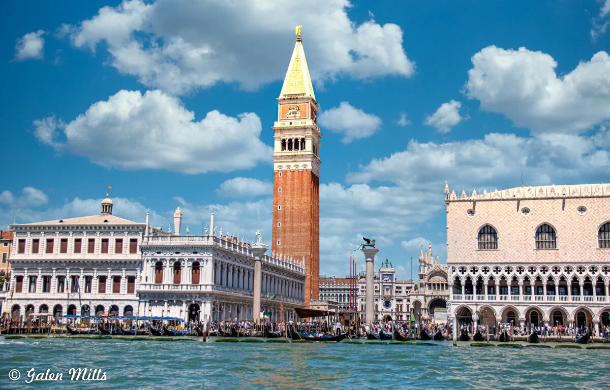 Venice skyline with St. Mark's Campanile and Doge's Palace seen from the water, featuring historic architecture against a blue sky with clouds.