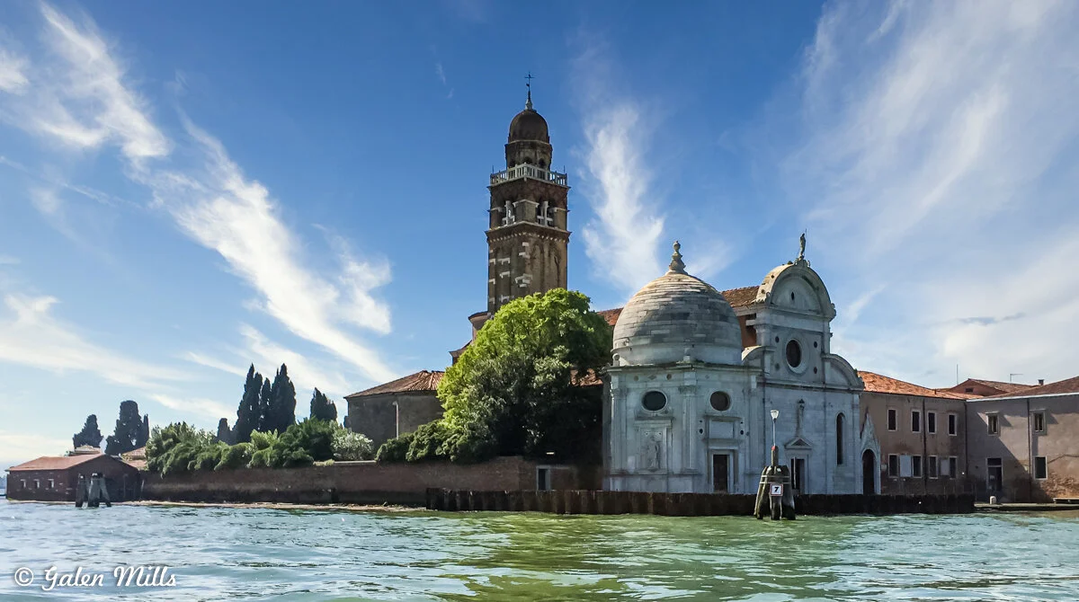 View of San Michele island in Venice, Italy, featuring the prominent Church of San Michele with its dome and bell tower, surrounded by greenery and calm waters under a clear blue sky.