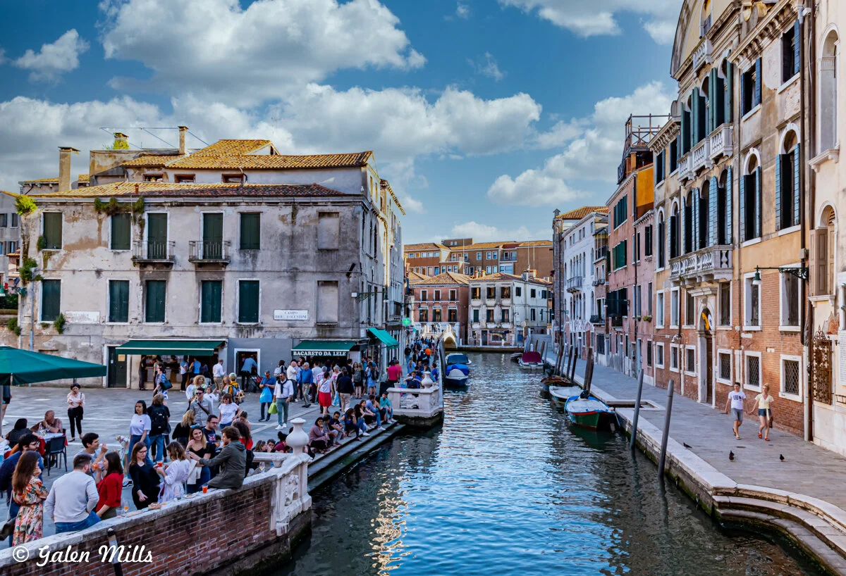 People gathered along a canal in Venice, Italy, with historic buildings and boats docked at the sides, under a blue sky with clouds.