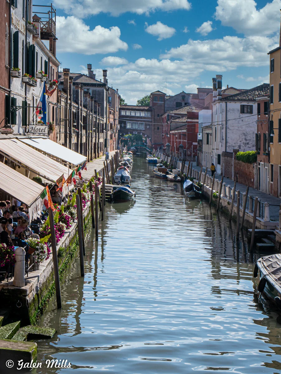 Venetian canal with boats, lined by old buildings, outdoor dining areas, and flower decorations under a blue sky with clouds.