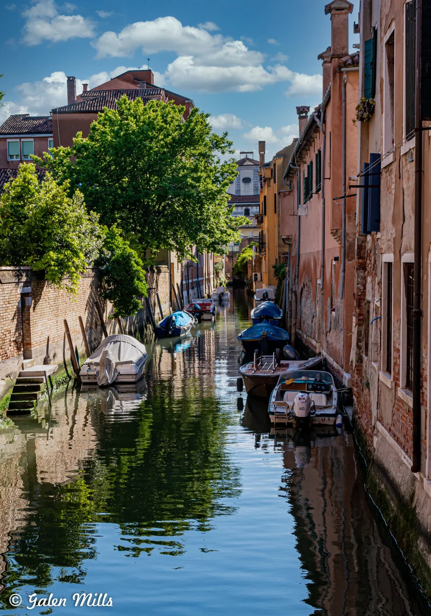 Narrow canal in Venice, lined with old buildings and small boats, bordered by trees and under a blue sky with clouds.