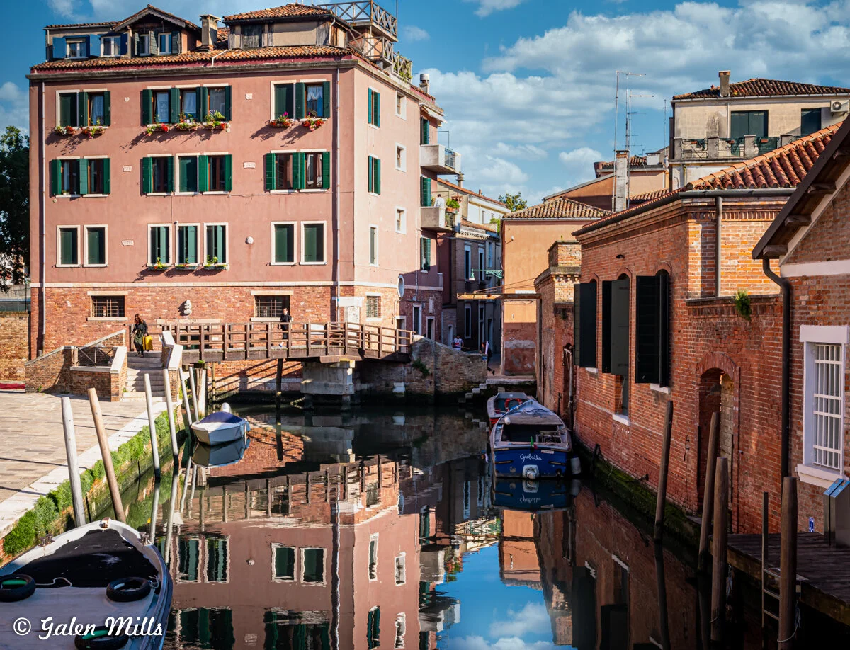 Scenic canal in Venice with colorful buildings, wooden bridge, boats, and reflections in water on a sunny day.