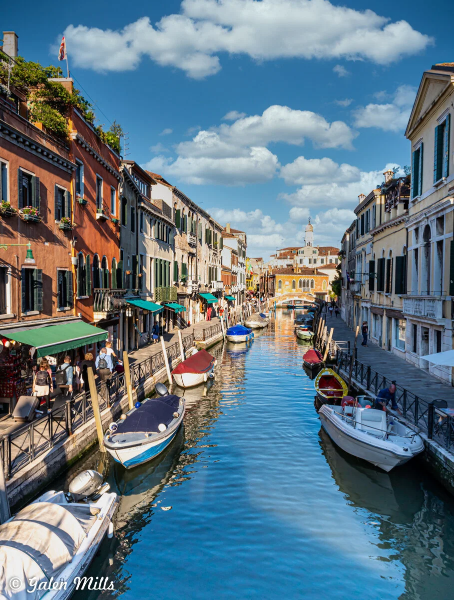 Scenic canal in Venice with boats, colorful buildings, and blue sky with clouds.