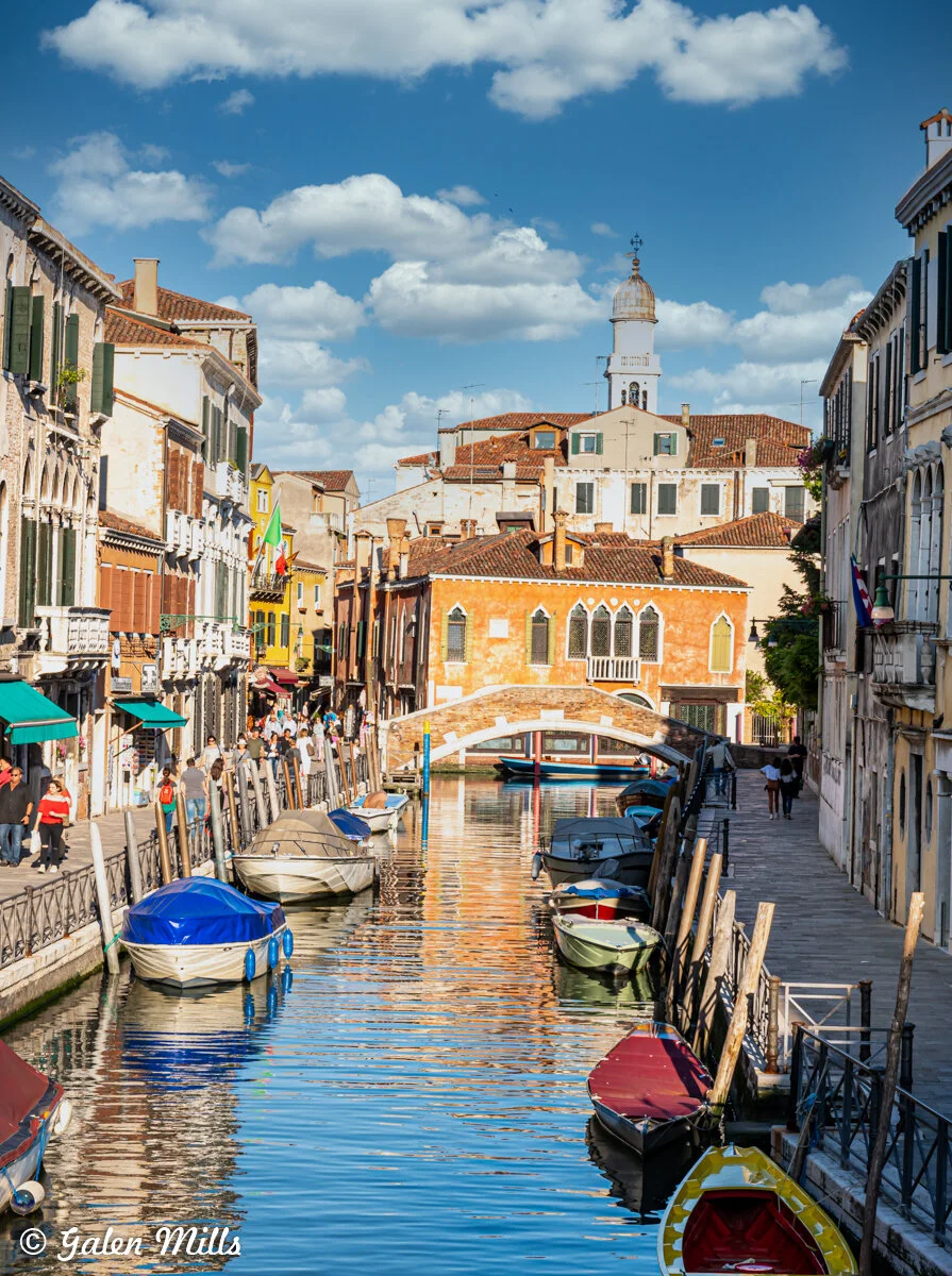 Venice canal with boats and colorful buildings, featuring a bridge and a tower in the background under a partly cloudy sky.